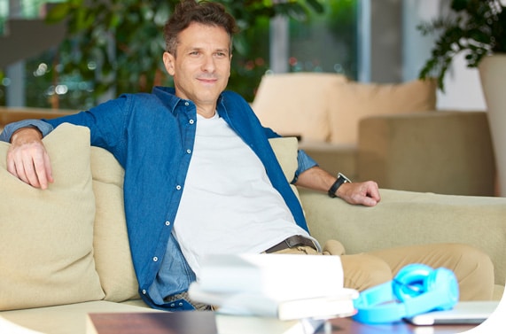HR Business A man wearing a blue shirt over a white t-shirt sits in a casual corporate environment on a beige sofa and smiles